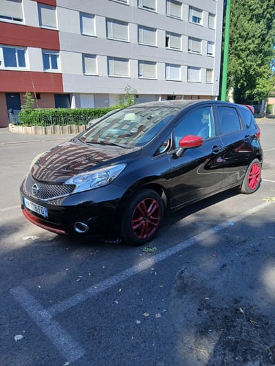 A black Nissan Note car with red accents on the side mirrors, front bumper, and wheels is parked in a paved lot in front of a multi-story apartment building. The car's license plate reads "HH 776 FD 08". The apartment building has a facade with sections of red and light gray, and numerous windows with closed white shutters. Green foliage and trees are visible behind the building and to the right. The parking lot surface is asphalt with white painted parking lines. The scene appears to be a residential area in the city of Charleville-Mézières, France, likely during daylight. There are no people or animals visible in the image.