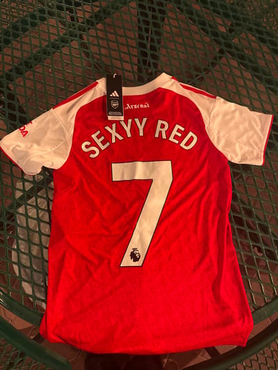 The image shows an Arsenal soccer jersey on a green metal grate table, likely outdoors. The jersey is predominantly red with white sleeves and shoulders. On the back, the white number "7" is prominently displayed, with the words "SEXY RED" above it. A black Adidas tag hangs from the jersey's collar. The environment appears to be a patio or outdoor seating area. While the specific location cannot be determined from the image, it is claimed to be in Indian Wells, USA. No people are visible in the shot.