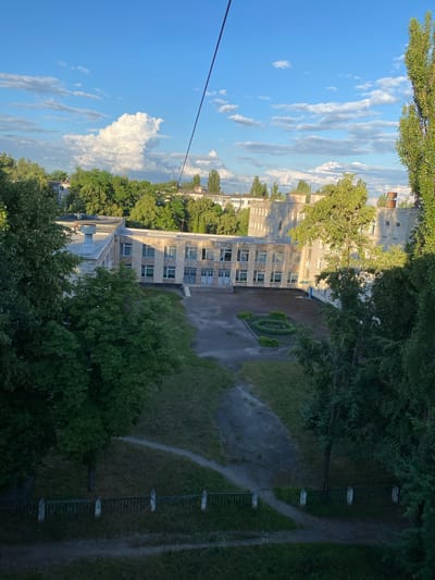 This media file captures an outdoor scene, likely in Shostka, Ukraine, during the daytime. The perspective is elevated, looking down on a building complex surrounded by lush greenery.
The main subjects are a multi-story building with a light-colored facade, punctuated by numerous windows. The building appears to be of institutional or residential design, with a somewhat dated architectural style. In front of the building, a paved area leads to a circular garden bed of neatly trimmed bushes. Beyond this, grassy areas and more trees are visible. To the right, a pathway winds through the grass, bordered by a metal fence with spaced supports. The surrounding environment is dominated by mature trees, their green foliage creating a dense canopy in places.
The time of day appears to be late afternoon or early evening, suggested by the warm, slightly diffused sunlight casting shadows and illuminating the clouds in the sky. The weather seems pleasant, with a clear blue sky dotted with fluffy white and wispy gray clouds.
Notable details include a dark wire or cable running diagonally across the upper left portion of the image, possibly from a power line or other overhead infrastructure. The overall impression is one of a quiet, established community or institution within a natural setting. There is no visible text in the image.