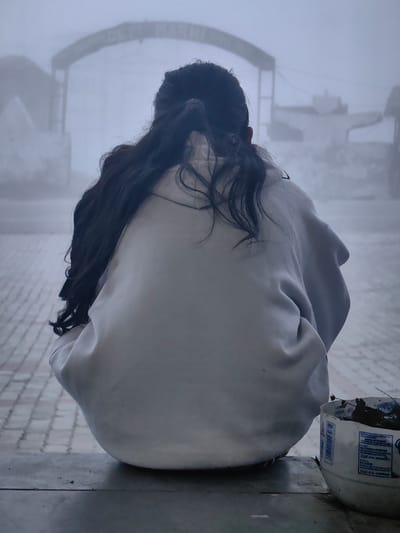 A person with long, dark hair is sitting with their back to the camera, facing an archway that appears to be the entrance to Marhi, India. The person is wearing a light-colored, loose-fitting garment, possibly a sweatshirt. They are crouched down on a tiled surface. The environment is shrouded in fog, obscuring much of the background and creating a muted, atmospheric scene. In the lower right corner, a white plastic container filled with leaves and debris is visible. The overall impression is one of solitude and quiet contemplation in a foggy, possibly cold, setting.