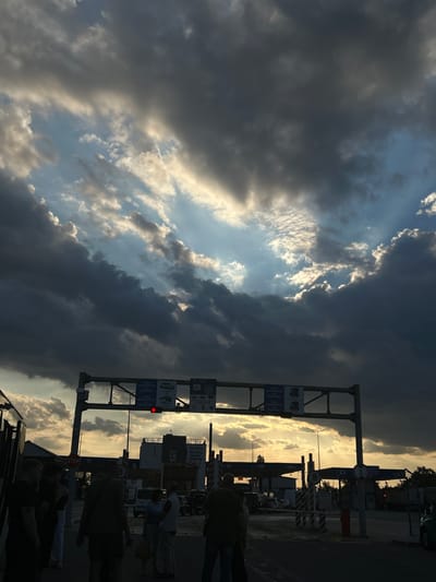 The image depicts a scene at the Palanca border crossing in Moldova, during what appears to be sunset. The sky is dramatic, with heavy, dark clouds interspersed with rays of sunlight breaking through, creating a striking contrast. In the foreground, silhouetted figures of people are visible, some standing and observing, while others appear to be in transit. A large gantry structure, characteristic of border control points, spans across the road, displaying signage in what appears to be Romanian and English. A red traffic light is illuminated on this structure. Various vehicles, including what looks like a bus on the left, are present, suggesting ongoing traffic flow. The overall atmosphere is somber due to the lighting conditions, but also dynamic due to the weather and the presence of people and infrastructure at a border crossing. The background features buildings and trees, consistent with a border town or a transportation hub.