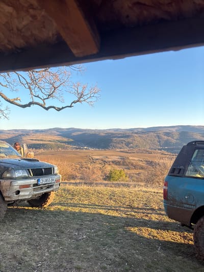 The scene depicts an outdoor landscape overlooking the village of Baylovo, Bulgaria, under a clear blue sky. The viewpoint is from beneath a wooden overhang structure, partially obscuring the top of the frame. In the immediate foreground, two mud-splattered off-road vehicles are parked on a dry, grassy hillside.
On the left, a dark green Mitsubishi Pajero Sport SUV, heavily coated with mud, presents its front left quarter. The Bulgarian license plate "CB 0855 BK" is affixed to the front bumper. A black snorkel is visible on the passenger side A-pillar. The front passenger door is open, and a male individual wearing a camouflage-patterned jacket is partially visible, leaning into or interacting with the vehicle's interior.
On the right, the rear quarter of a turquoise or light blue SUV, also heavily covered in mud, is partially visible. A large, muddy off-road tire is present.
Beyond the vehicles, a valley unfolds into the distance. The terrain consists of dry, light brown fields and sparse, leafless deciduous vegetation. Winding dirt tracks traverse the landscape. In the mid-ground, the village of Baylovo is visible, characterized by clusters of buildings amidst the valley floor, framed by undulating, forested hills in the far background. A large, bare tree branch extends from the left side of the frame into the clear, cloudless blue sky.