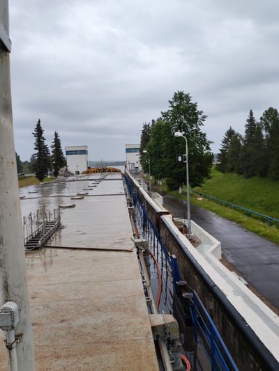 The image shows a view from a high vantage point of a lock system, likely part of a waterway or canal, on an overcast day. In the foreground, to the left, is a portion of a white, cylindrical structure, possibly a support pillar or part of a building. To the right of this pillar, a large, flat, wet concrete surface extends into the midground, marked with horizontal lines and several circular, raised elements that appear to be hatches or covers. A metal staircase with railings leads down from this surface towards the left. Further into the scene, the concrete surface appears to transition into a lock chamber lined with a dark, thick material along its edge, with a blue metal railing running along the top.
Across the lock chamber, on the other side of the water (which is not directly visible), are two identical, multi-story white buildings with blue-framed windows, situated behind rows of tall, dark green evergreen trees. These buildings have flat roofs and appear to be control or operational structures for the lock. Between the buildings and further in the background, more trees and a body of water are visible, along with a cloudy sky.
A paved road runs parallel to the lock chamber on the right side of the image, bordered by a grassy slope and more trees. A lamppost with a sign displaying the number "6" is visible along this road. The overall environment suggests an industrial or engineering facility, likely a dam or lock for water management, in a semi-rural or forested area. No people are visible in the image. The presence of water on the surfaces indicates recent rain or damp conditions. The location is identified as Belousovo, Russia.