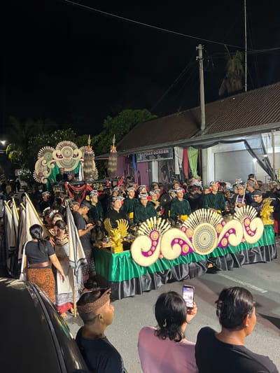 A nighttime street scene in Munggu, Indonesia, depicts a cultural or religious procession. The primary focus is a large, elaborately decorated float moving along an asphalt road. This float features a base draped in green and black fabric, adorned with intricate, light-colored, fan-like and swirling patterns, and two golden Garuda figures flanking small gongs. Behind it, a second, taller float displays similar decorative patterns and two prominent tall, dark, textured ornaments.
Approximately 30-40 individuals participate in the procession. Many wear uniform dark green long-sleeved shirts, black trousers, and traditional Balinese Udeng (head wraps). Some are positioned behind the main float, appearing to carry or attend to instruments. Others walk alongside, carrying white flags with black patterned edges on staffs.
In the foreground, several spectators observe the event from the side of the street. One individual, seen from behind in a pink shirt, holds a smartphone vertically, recording the procession. Another spectator in a black shirt and traditional headwear is positioned to their left. Part of a dark-colored vehicle is visible in the lower left corner.
The environment includes buildings lining the street, such as a structure with a brown tiled roof and a visible sign for "AFDAN GORDEN." A utility pole with power lines extends above the street. Artificial lighting illuminates the scene against a dark night sky. Palm trees and other foliage are visible in the background.