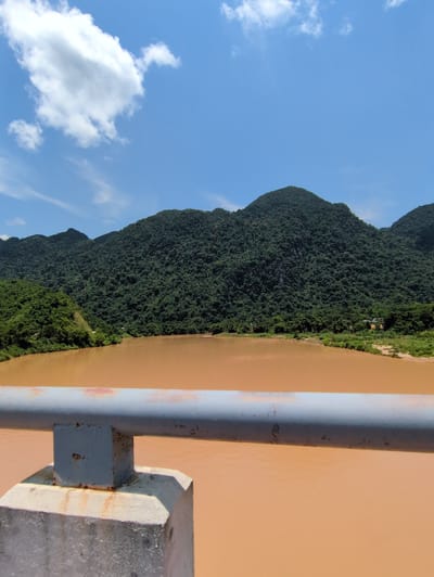 The image shows a wide, brown river in front of a backdrop of lush green mountains. The river takes up the lower half of the frame, its surface appearing calm with slight ripples. The mountains in the background are densely covered with trees, with peaks and ridges creating a dramatic silhouette against a bright blue sky with scattered white clouds. In the foreground, a concrete structure with a weathered metal railing is visible, suggesting the image was taken from a bridge or elevated viewpoint. The scene appears to be a natural landscape, likely in a rural or undeveloped area. The location is Hồi Xuân Commune, Vietnam.