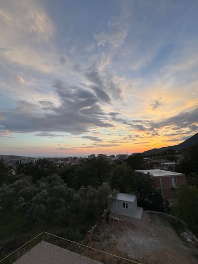 The image depicts a sunset over Burtaiši, Montenegro. The sky is filled with a mixture of clouds, some dark gray and others illuminated by the setting sun in hues of orange, yellow, and pink. In the foreground, there is a paved area with a metal railing, suggesting a balcony or viewing platform. Beyond this, a dense cluster of green trees and vegetation covers the hillside.

Further down the slope, there are a few buildings. One is a small, gray, prefabricated structure with two windows and a concrete base. To its right, a larger, unfinished brick building with a concrete roof and visible window frames is situated. Beyond these structures, the cityscape of Burtaiši stretches towards the horizon, with numerous residential buildings visible. The sea is visible in the distance, reflecting the colors of the sunset. A mountain range is visible on the right side of the image, its silhouette dark against the vibrant sky. The overall scene captures a tranquil evening view of the coastal town.