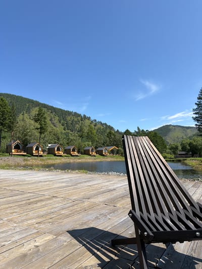 A scenic view of a resort area with small wooden cabins situated along the bank of a tranquil lake, with a lush, green, tree-covered mountainside in the background. The foreground is dominated by a wooden deck, upon which rests a dark wooden lounger chair, casting a patterned shadow across the weathered planks. The sky is a clear, vibrant blue with a few wispy white clouds. The cabins, each with a triangular roof and large glass front, appear unoccupied. The water of the lake is calm and reflects the surrounding landscape. The overall impression is one of peaceful, natural beauty, likely a tourist destination.