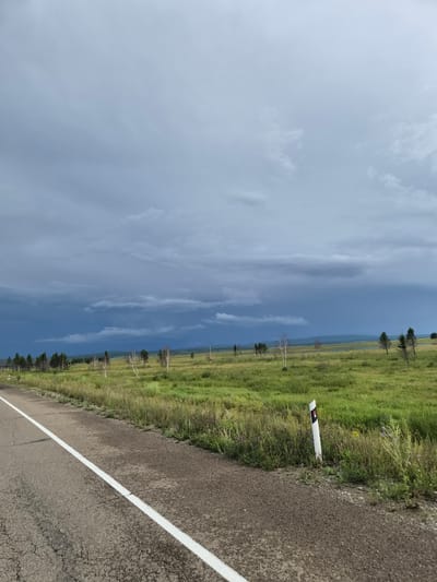 A paved road with a white line marking runs along the left side of the frame, bordering a grassy field. Scattered trees, some leafless and white-barked, are visible in the distance across the field. The sky is overcast with dark, heavy clouds, suggesting impending rain, and a sliver of blue sky is visible on the horizon. A white road marker with red and black markings stands in the grass near the road. The landscape is flat and appears rural, possibly on the outskirts of Mukhor-Konduy, Russia. There are no people or animals visible in the image.