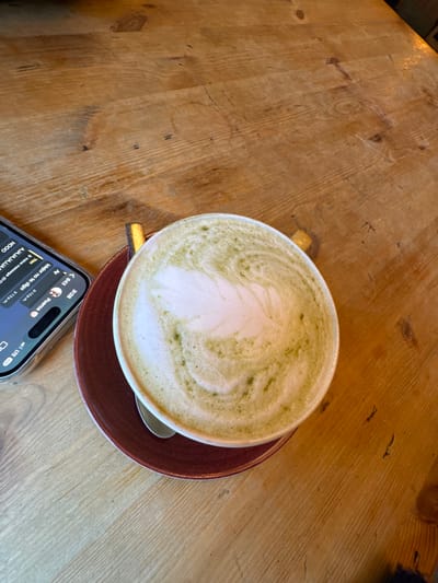 A bird's-eye view shows a matcha latte with a creamy white leaf-like design on a wooden table. The latte sits on a brown saucer with a spoon. A phone rests near the bottom, displaying a chat message thread. The phone's status bar includes the time, signal strength, and battery information. The table's wood grain is visible and adds a rustic feel to the image. The overall aesthetic is casual and contemporary.