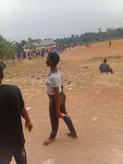 An outdoor gathering is captured in this shot, where a crowd gathers on the edge of a field. In the foreground, a person clad in a white shirt and dark trousers holds a red object, perhaps a bottle or container, while another individual in black is partly visible. The expansive field appears dry, with scattered patches of grass. In the distance, more people are gathered, and some figures are seen on the field. The overall scene conveys a communal or event atmosphere.