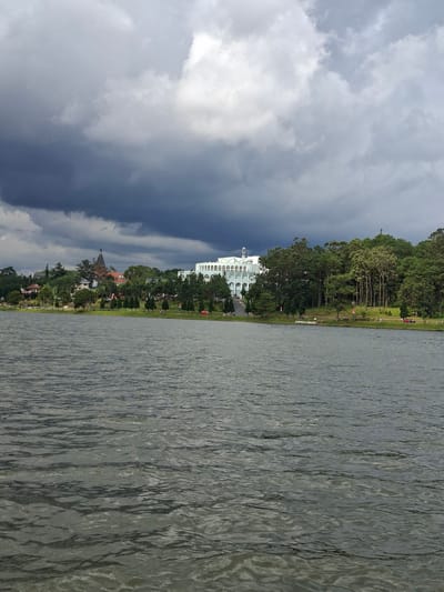 The image captures a serene view of Xuan Huong Lake in Da Lat, Vietnam, on a cloudy day. The foreground is dominated by the rippling surface of the dark, calm water. In the midground, a lush green shoreline is dotted with various trees, including tall pines. Beyond the trees, several buildings are visible. A large, white building with a distinctive arched facade stands prominently on the right. To the left, a darker, more traditional-looking building with a conical tower can be seen, nestled among the trees and hills. The background is filled with a dramatic sky, with heavy, dark storm clouds gathering above, contrasting with patches of lighter sky. Several individuals are scattered along the shoreline, some sitting on benches or walking, while a red car is parked near the edge of the lake. The overall atmosphere is one of peaceful contemplation amidst a slightly overcast, natural setting.