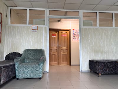 The image depicts a waiting area with two upholstered benches and an armchair. The walls are covered in a textured, light-colored wallpaper. A doorway in the center leads to a hallway with two wooden doors. On the left door, a red sign reads "Гараж" (Garage). On the right door, a sign in a red square reads "ДЕРЖАТЬ ЗАКРЫТОЙ" (Keep closed). To the right of the doorway, a red poster with images and text in Russian is displayed, titled "УГОЛОК ПО ОХРАН" (Corner for safety). The floor is tiled in a neutral color. The lighting suggests it is daytime, and the indoor setting does not provide weather cues. The scene appears to be a public building, possibly an office, industrial facility, or community center, in Polarnie Zori, Russia. There are no people visible in the image.