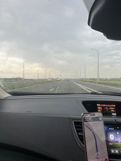 The image, taken from inside a car driving on the E40 highway, shows a view through a rain-streaked windshield. A navigation app on a phone, mounted on the dashboard, indicates the vehicle is near Veurne, Belgium, and approximately 120 kilometers from the E19 highway. The car's dashboard display shows a time of 18:00, an average MPG of 19.7, and a speed of 25 mph. The road outside is wet, with a line of streetlights visible in the distance.
