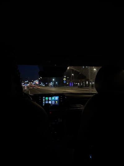 A view from inside a car at night, looking forward through the windshield. The car is in motion on a city street, with traffic and streetlights visible ahead. The dashboard of the car is illuminated, showing a navigation screen with icons for various apps. There are two people in the car, their silhouetted heads visible in the foreground. The windshield is wet with rain. The scene appears to be in a major urban area with multi-lane roads and commercial buildings lining the street.
