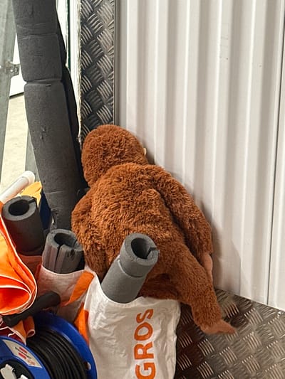 The image shows a close-up of items stored in a vehicle or storage area. A brown stuffed animal, possibly a monkey or bear, lies amongst various objects. These include grey pipe insulation tubes, a white bag with the word "GROS" printed in orange, a partially visible orange fabric item, and a blue reel of black cable. The background consists of textured metal paneling and a white corrugated surface. The image does not provide enough information to confirm a specific location in Volketswil, Switzerland. The scene shows storage of materials and possibly personal items within a vehicle or storage space.