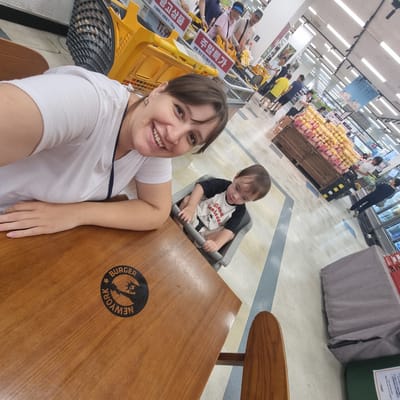 In a brightly lit supermarket in Wonju-si, South Korea, a woman in a white t-shirt smiles at the camera, her left arm resting on a wooden table. To her right, a toddler sits in a high chair, wearing a black and white shirt with a graphic. The table has a circular black logo with the text "BURGER NEW YORK". In the background, other shoppers and grocery carts are visible, with displays of produce and shelves stocked with goods. The floor has a patterned tile design, and the ceiling is lined with fluorescent lights.