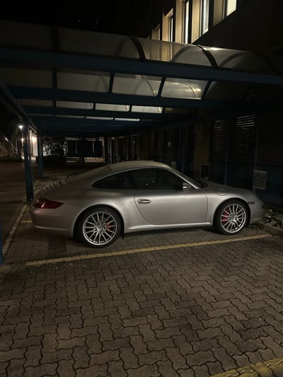 The image depicts a silver Porsche parked in a parking space delineated by yellow lines in Wetzikon (ZH), Switzerland. The vehicle is situated under a structure with blue support beams and a curved, translucent roof, possibly a walkway. The ground is paved with interlocking gray bricks. Additional architectural details can be observed in the background, including a building structure with visible windows and an exterior lighting fixture. The scene appears to be captured at night.