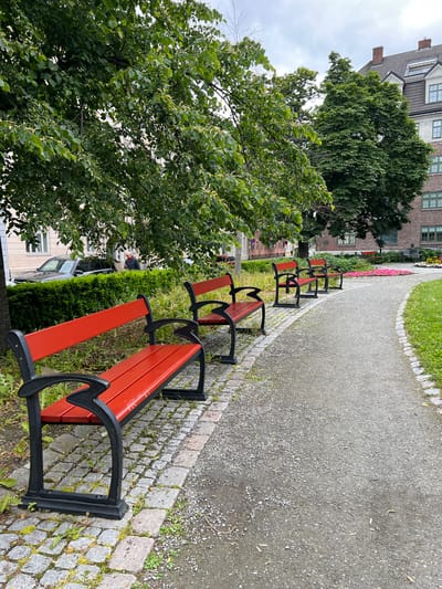 This image depicts a park scene in Trondheim, Norway. In the foreground, a paved pathway with a gravel surface curves to the right. Along the left edge of the pathway are several red park benches with black metal frames. The benches are arranged in a row, with the closest bench in sharp focus and others receding into the distance. The pathway is bordered by cobblestone edging on the left and grass on the right. Beyond the benches, a lush green tree canopy overhangs the area, with sunlight filtering through the leaves. In the background, several buildings are visible, including a multi-story brick building with red-tiled roofs and chimneys, typical of Trondheim's architecture. A person in a red jacket is visible standing near a hedge and a parked black car in the mid-ground, to the left of the pathway. Further into the park, there appears to be a flowerbed with pink flowers and more park benches, one of which is occupied by a seated figure. The sky is overcast, suggesting a cloudy day.