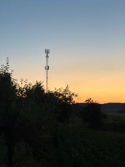The image shows a silhouette of a cell tower against a gradient sky of pale blue transitioning to orange. In the foreground, dark silhouettes of trees and foliage obscure the lower half of the scene. The cell tower, a slender metal structure with multiple antenna arrays at its peak, stands tall in the middle-right portion of the frame. Beyond the foliage, a dark, hilly landscape is visible, with hints of distant structures and a fence line. The overall scene suggests a rural or semi-rural setting during twilight or dawn. The location is identified as Радашковіцкі сельскі Савет, Belarus. No people or specific actions are depicted.