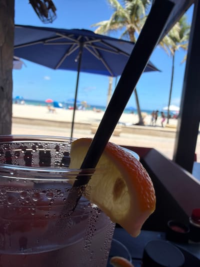 A close-up shot shows a beverage in a glass with a slice of orange and a black straw. The background depicts a beach scene in Hollywood, Florida, with palm trees, beach umbrellas, and people walking on the sand. The ocean is visible in the distance under a clear blue sky.