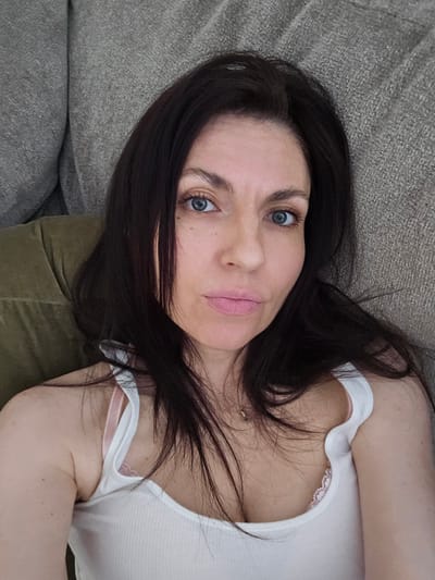 The image is a selfie of a woman with dark hair and blue eyes, lying on a couch. She is wearing a white tank top. The background appears to be a living room with a textured couch. The location is identified as Liberty Hill, United States of America.