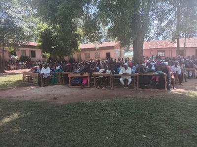 An outdoor public gathering is depicted in Idakho South ward, Kenya. Approximately 150-200 individuals, primarily adults, are assembled in a dirt clearing featuring patches of grass and shaded by multiple large trees. Numerous people are seated on wooden school desks and benches arranged in rows, facing an unseen central point. Additional individuals are visible standing or seated further back near several single-story buildings with light-colored walls and corrugated iron roofs, likely part of an institutional complex. The scene is illuminated by strong natural daylight.