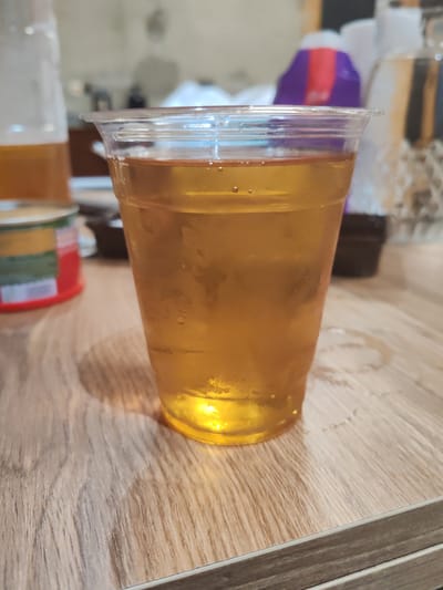 This media file is a close-up, eye-level shot of a clear plastic cup filled with a golden, amber-colored liquid, likely beer or a similar beverage. The cup is condensation-covered, suggesting it is cold. Several small bubbles are visible within the liquid. The cup sits on a light-colored wooden surface, possibly a table. To the left of the cup, partially visible, is a glass bottle also containing a similar amber liquid, and a red and green cylindrical object. In the background, out of focus, are various objects including what appears to be a purple and red bag, and other indistinct shapes. The lighting suggests it is likely indoors, possibly in the evening or under artificial light, as there are no clear indicators of natural light or outdoor weather. The scene appears casual and relaxed, with the focus solely on the drink. No visible text is clearly discernible. The setting is implied to be Ortabatumi, Georgia, but this context is not visually evident within the image itself.