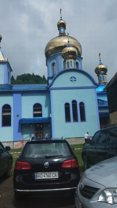 This image depicts the exterior of a bright blue church with golden domes. The building features arched windows and decorative blue trim. Several cars are parked in front of the church. A black Volkswagen is visible in the foreground with a license plate reading "AO 4160 CA". The scene appears to be in Kvasy, Ukraine, indicated by the context. The sky is overcast, suggesting a cloudy day. A person in a white shirt is standing near one of the parked cars. The overall impression is that of a community building in a somewhat rural or suburban setting. The church's vibrant colors stand out against the muted sky.