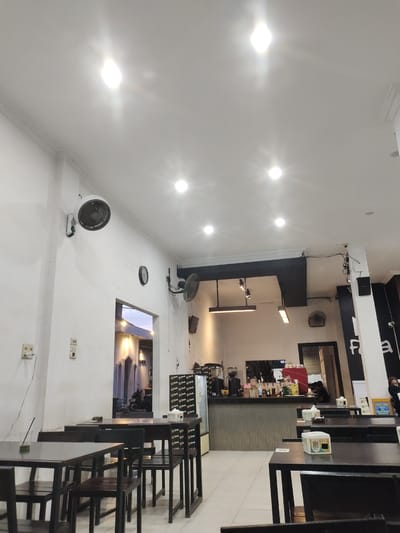 The image depicts the interior of a cafe in Lhoksukon, Indonesia, as indicated by the text on a sign. The perspective is from a low angle, looking upwards and across the space. The cafe features dark wooden tables and chairs arranged in rows. In the foreground, several tables and chairs are visible, with one table in the lower left having a glass with a straw and a green liquid.
The walls are painted white, and the ceiling is white with recessed lighting, some of which are very bright, creating glare. Mounted on the wall is a white oscillating fan. A black analog clock is also visible on the wall. To the right of the clock, another black oscillating fan is mounted lower down. Further to the right, a tall white pillar supports part of the ceiling structure, and on this pillar, a black speaker and some wiring are attached.
A doorway or opening in the wall leads to another part of the building, where a motorcycle is parked and architectural elements with arched windows are visible. Beyond this, the exterior light suggests it might be dusk or early evening.
The main counter area of the cafe is visible in the mid-ground. It has a ribbed metal front and is topped with a dark surface where coffee machines and various bottles of syrups or sauces are placed. A person is partially visible behind the counter, appearing to be working.
On the tables, there are small white boxes, possibly for sugar packets or napkins, and some paper placemats. Signs with text and graphics are visible on some surfaces. In the background, towards the right edge, a person is seated at a table. The floor is tiled with light-colored square tiles. The overall impression is of a modestly appointed, functional cafe.