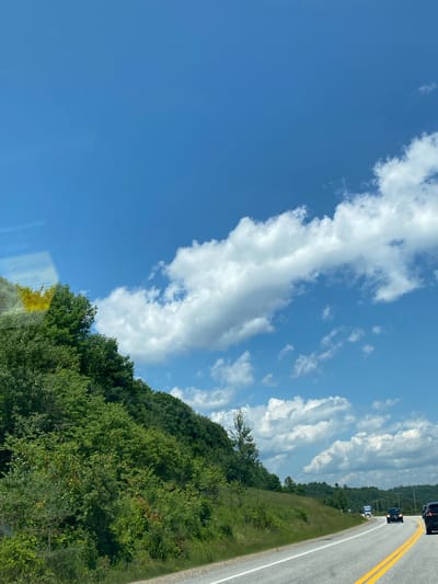 The image shows a bright, sunny day with a clear blue sky filled with fluffy white clouds. A winding road, marked with double yellow lines and a white lane divider, curves through a landscape dominated by lush green trees and vegetation. The road appears to be a highway, with a few vehicles visible, including a black SUV and a truck. The terrain is hilly, with a steep, tree-covered incline on the left side of the road and rolling hills in the distance. The scene suggests a rural or suburban setting, possibly a scenic route. There is no discernible location information in the image to place it in Ripon, Canada.