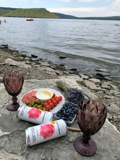The image displays a detailed picnic scene situated on a rocky shoreline in Horaivka, Ukraine. In the immediate foreground, a cluster of items is arranged on flat, greyish-brown rock formations. These items include two dark purple, patterned glass goblets positioned on the left and right sides. Centrally, a white scalloped plate holds an assortment of food: slices of reddish sausage or salami, small green pickles, and a pile of red cherry tomatoes. A small white bowl on the plate contains a yellowish, creamy substance. Directly in front of the plate are two white aluminum cans, identified as "spraga" kombucha with cherry illustrations. To the right of the plate, a clear plastic container is filled with blueberries. Beside the blueberries, a small, tied bundle of dried purple lavender flowers is visible, alongside a small bundle of dried straw or grass.
The midground transitions to the water's edge, where small waves lap against the stony shore, which is composed of numerous flat, grey rocks. The background features a large body of water, consistent with the Dniester River, extending into the distance under a partly cloudy sky. Several individuals are observed swimming or wading in the water. A red motorboat with a visible wake is traversing the water from left to right in the mid-distance. Across the water, distant green, rolling hills are prominently visible.