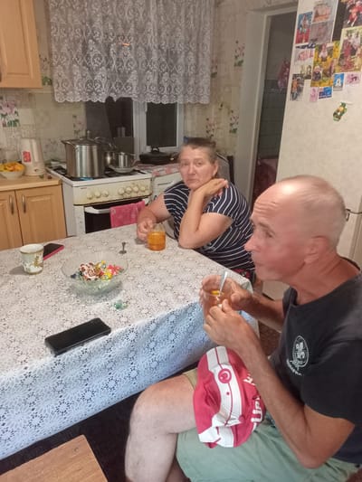 In Shabo, Ukraine, two individuals are seated at a table covered with a white, lace-patterned tablecloth in what appears to be a kitchen. On the left, a woman with graying hair, wearing a striped shirt, is looking towards the camera. In the foreground, a bald man with light skin, wearing a dark t-shirt with a logo, is facing to the right and holding a glass with a beverage and a spoon.
On the table, a glass bowl filled with wrapped candies is centrally located. To the left of the bowl is a decorative cup with gold accents, and next to it, a black mobile phone. A second black mobile phone is visible further down the table. A drink in a glass with ice and a spoon is in front of the man.
In the background, a white gas stove with silver pots is visible. A white electric kettle is on the counter to the left of the stove, next to a bowl of oranges. Above the stove, a white lace curtain hangs across a window. The walls are decorated with a floral wallpaper pattern and numerous photos are affixed to a vertical surface on the right, suggesting it may be a refrigerator. A red and white cap with the text "TURKIYE" and a crescent moon symbol is resting on the man's lap.