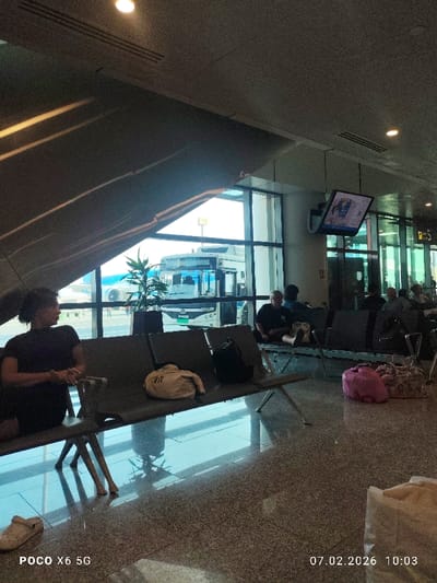 An airport waiting area shows passengers seated on metal benches. Luggage sits next to them on the floor or on the benches. Large windows reveal a bus outside and a glimpse of an airplane. A television screen hangs from the ceiling. A potted plant sits next to the windows. The floor is shiny and reflects the light. A pair of white clogs sits near the corner of the image. A digital watermark shows the device and date.