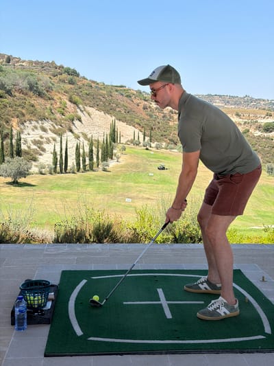 A man is practicing golf on a driving range in Tsada, Cyprus. He is standing on a green mat with white lines, holding a golf club and is about to hit a golf ball. There are several golf balls in a green basket next to a bottle of water. In the background, there is a green golf course with hills, trees, and a golf cart. The sky is clear and blue. The man is wearing a green polo shirt, brown shorts, a cap, sunglasses, and sneakers.