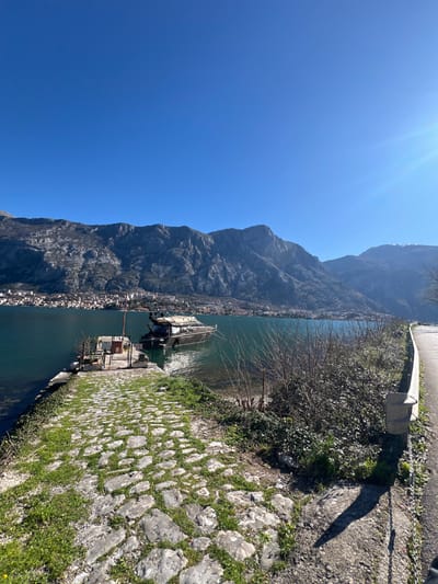 The image depicts a sunlit coastal scene captured in Bogdašići, Montenegro, featuring a body of water bordered by a mountainous landscape. In the left foreground, a stone-paved path, partially covered with green grass, descends toward the water. This path terminates at a small, rectangular concrete pier where a dark-hulled motor yacht, equipped with a light-colored canopy, is moored. To the right of the paved path, a strip of sparse vegetation and bare ground runs parallel to the water, adjacent to a paved road. This road is delineated by a silver metal guardrail on its left edge. Across the calm, greenish-blue water, a densely settled town with multiple buildings is visible along the distant shoreline. Behind the town, a prominent range of rugged, grey mountains rises steeply, dominating the background. The sky above is uniformly clear and bright blue. Direct sunlight from the upper right generates a visible lens flare and casts distinct shadows on the foreground elements. No individuals are present in the scene.