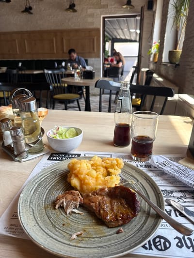 The image depicts a close-up view of a table in what appears to be a restaurant. In the foreground, a plate holds a meal consisting of mashed potatoes and a piece of meat, with a fork resting on the side. To the left of the plate are salt and pepper shakers and a bottle of oil and vinegar. A small bowl of salad, a glass of dark beverage, and a bottle of cola are also visible on the table. The background shows other tables, chairs, and patrons, suggesting a lively dining atmosphere. The restaurant's interior features wooden panels, brick walls, and hanging lamps. The overall impression is one of a casual dining experience.