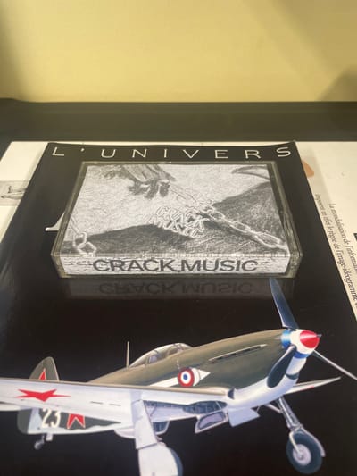 This image captures an indoor scene in Reventin-Vaugris, France, displaying several objects arranged on a dark, reflective surface.
A clear plastic CD jewel case, containing an item labeled "CRACK MUSIC," is positioned centrally. The CD cover art is a black and white illustration depicting two hands, one holding a heavy chain, set against a rough, textured background. The words "CRACK MUSIC" are visible vertically on the spine of the case and horizontally along the bottom edge of the front cover.
The CD case rests on an open publication. The visible left page is dark, featuring the partially cropped title "L'UNIVERS" in white capital letters across the top. The right page is lighter, displaying French text, including "La mondialisation de l'information..." An illustration of a propeller-driven aircraft, likely a military fighter, is prominent on the lower portion of the dark page, beneath the CD case. This aircraft is olive green with light underwings, features a four-bladed propeller with a red and blue spinner, and includes distinct markings: a red star with a white outline and the number "23" on the visible left wing, and a French tricolor roundel (concentric red, white, and blue circles) with a white forward-pointing arrow on the fuselage side near the cockpit.
The dark surface beneath the objects exhibits a faint reflection of the "CRACK MUSIC" text. Behind this arrangement, a plain, light yellow-beige wall occupies the upper portion of the frame, separated from the surface by a thin, dark horizontal line. No individuals are present in the scene.
