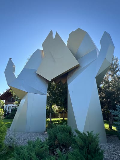 The image captures a large, abstract metallic sculpture with sharp, geometric angles. The sculpture is silver and gold in color and appears to be composed of several large, angular panels. It is situated outdoors in a well-maintained green space with trees and shrubs. To the left of the sculpture, a portion of a wooden building with a white facade and windows is visible, with a sign that reads "КОНН" above the entrance. The background features a clear blue sky, sunlight, and a variety of trees, including a tall evergreen tree on the right side. The ground around the sculpture is covered with small gray gravel, and lush green bushes are in the foreground. The location is identified as Plastunivska, Russia. There are no people visible in the image.