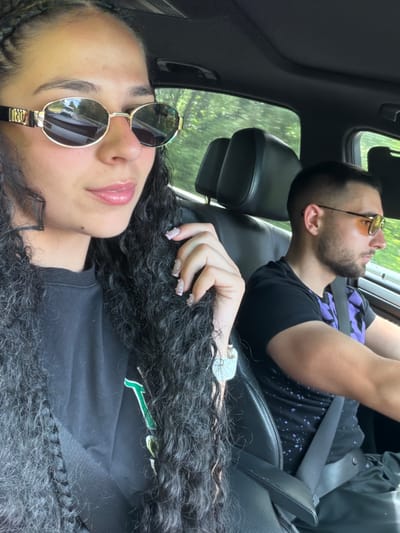 The image shows two individuals in a car, likely in the city of Rahovtsi, Bulgaria. The woman in the foreground is wearing large, oval, gold-rimmed sunglasses and has long, dark, curly hair braided in the front. She is looking towards the camera, and her lips are slightly parted, appearing to be wearing lip gloss. She is wearing a black t-shirt with a green and white logo visible on the left side of her chest. Her left hand is raised, with manicured nails, near her face.

The man in the background is driving the car. He is wearing orange-tinted sunglasses, a black t-shirt with a purple splatter pattern, and a seatbelt. His hair is short and neatly cut. The interior of the car is visible, including black leather seats and the car's roof. Through the car windows, trees and greenery are visible, suggesting a location with natural surroundings. The lighting indicates it is daytime, possibly sunny.
