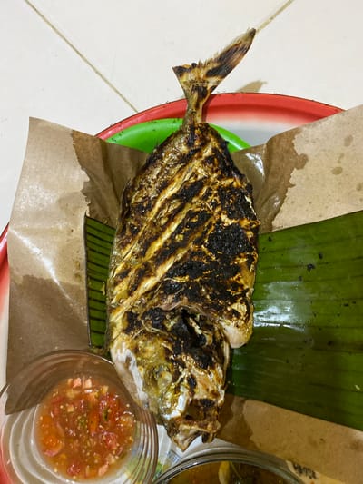 A high-angle, close-up photograph captures a whole grilled fish, likely a trevally, served as part of a meal. The fish, oriented horizontally with its tail pointing left, dominates the center of the frame. It displays distinct char marks across its skin, indicating it has been grilled, giving it a dark brown and golden-brown, somewhat glossy finish.
The fish rests on a bed composed of a large, dark green banana leaf and a piece of brown parchment paper. Both the leaf and paper are placed on a red and green plastic tray, which is largely obscured. The parchment paper shows signs of grease absorption, particularly around the fish.
To the lower right of the fish, a small, clear glass bowl contains a vibrant red salsa or sambal, featuring visible chunks of tomato, chili, and possibly onion in a liquid base. A portion of another bowl, holding a yellowish liquid that could be soup or another sauce, is visible in the upper right corner.
The setting appears to be a casual dining environment, possibly indoors or semi-outdoors, with the tray positioned on a light-colored tiled floor. Subtle details include a few small ants visible on the green part of the plastic plate near its red rim, and faint, partial, and illegible text on the rim of the glass bowl. The presentation style, using a banana leaf and a chili-based dipping sauce, is characteristic of Indonesian cuisine, aligning with the Tanjongkapai location context. No people are present, and the time of day is not discernible.