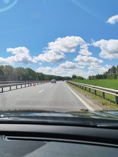 The image depicts a view from the driver's seat of a vehicle traveling on a highway. The road is paved with asphalt and marked with white lines. There are guardrails on both sides of the highway, with a green field and trees lining the right side and a dense forest on the left. The sky is blue with fluffy white clouds. Several cars are visible on the highway ahead. Orange traffic cones are placed on the left side of the road. The reflection of the sky and trees is visible on the dashboard. The location is identified as being in the city of Tatarstan, Russia, on a sunny day.