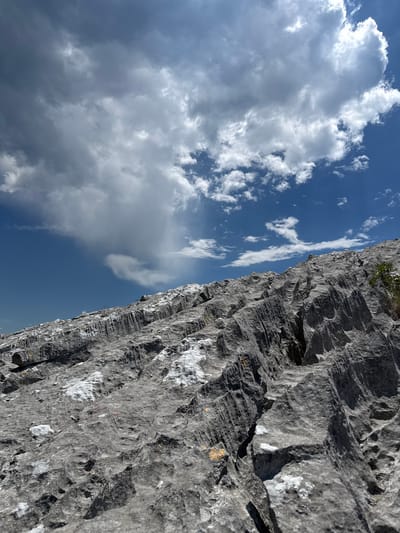 This is a close-up, vertical shot of a weathered limestone cliff face against a bright blue sky dotted with fluffy white clouds. The cliff occupies the right side of the frame, its textured surface revealing layers and fissures, with patches of white mineral deposits and hints of greenery at the very top. The left side of the image is dominated by the sky, where large cumulus clouds are illuminated by sunlight, casting soft shadows on their undersides. The composition creates a dramatic contrast between the rugged, solid rock and the vast, airy expanse of the sky. The lighting suggests it is daytime, likely midday given the brightness of the sun and the stark shadows on the rock. The scene evokes a sense of scale and natural grandeur, typical of a mountainous or coastal landscape. The location context of Žlijebi, Montenegro, suggests this is likely a view from a cliffside in that region, possibly a hiking trail or a climbing area. There is no visible text in the image.