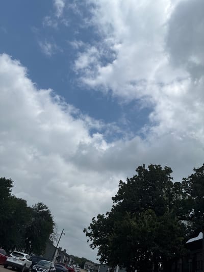 An outdoor, eye-level, medium shot captures a view of the sky, trees, and parked cars. The sky is partially cloudy with patches of blue visible. In the foreground, the tops of several large, dark green trees are prominent, their branches silhouetted against the sky. Below the trees, a row of parked cars is visible, including a white SUV, a black sedan, and several other vehicles. In the background, buildings that appear to be apartments or townhouses line a street, with a utility pole and wires also present. The overall scene suggests a typical residential or urban environment. The location is the city of Balch Springs, United States of America.