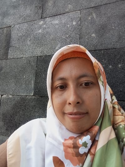A medium close-up image features a single adult female looking directly at the camera with a slight smile. She has dark eyes and medium-toned skin. She is wearing a white hijab with a prominent floral pattern in shades of orange, brown, and green. An orange, textured under-scarf is visible beneath the white hijab at her forehead. A small red pin is affixed to the right side of her hijab. The background consists of a dark gray wall constructed from rectangular, textured stone or tile blocks with visible grout lines. The specific location within Pasuruan, Indonesia, is not discernible from the visual information provided in the image.