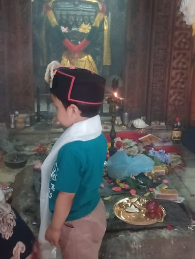 A young boy, facing away from the camera, stands in what appears to be a temple or shrine. He is wearing a black cap with red trim and a white scarf draped over his shoulders, over a teal t-shirt and tan pants. The boy is looking towards an altar area that is adorned with offerings.
The altar is cluttered with various items, including flowers (some real, some artificial), small boxes, a conch shell, and what looks like a bottle of dark liquid with a yellow label. A small oil lamp with a flame is visible, suggesting a religious ceremony or ritual is taking place, likely in the evening or a dimly lit interior. Petals are scattered on the floor around the altar.
Behind the altar, a dark, imposing structure with multiple carved faces or figures can be seen, likely a deity statue or religious iconography. The background also features wooden paneling with intricate carvings. The overall atmosphere seems reverent, with the focus on the boy and his interaction with the sacred space and its offerings.
The scene is set in Gojra, India. There is no visible text in the image that can be read clearly. The lighting suggests it is either dusk or an indoor setting with artificial light sources.