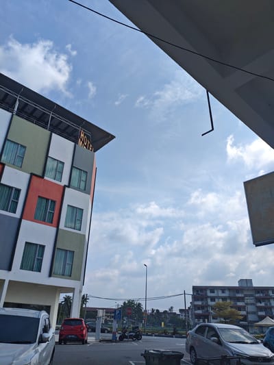 This is an exterior, eye-level, medium shot of a colorful hotel building on a sunny day in Port Dickson, Malaysia. The hotel has a modern design with geometric panels in white, grey, green, and red, each featuring windows with green curtains. A sign at the top of the building reads "HOTEL M...". In the foreground, parts of two cars are visible, a silver SUV on the left and a red sedan parked behind it. The red sedan has a license plate that reads "QRD 5692". A motorcycle is parked nearby. In the background, there are other buildings, including apartment complexes, streetlights, palm trees, and a stop sign. The sky is blue with scattered white clouds. The scene appears to be a parking lot area in front of the hotel.