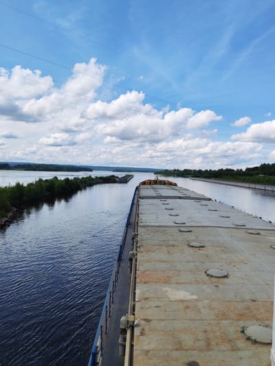 The image shows a view from the deck of a barge traveling along a calm, dark blue river. The barge's deck is metallic gray, covered in many circular hatches. On the left side of the river, there is a narrow strip of green trees and some rocky shoreline. In the distance, across the river, a treed landscape stretches to the horizon, meeting a bright blue sky with scattered white clouds. Power lines are visible overhead. The barge is moving towards a lock or a similar structure, indicated by a series of barges ahead. The location is the White Sea-Baltic Canal near the village of Anchimo in Russia.