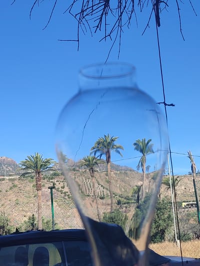 The image depicts a sun-drenched outdoor scene, possibly in Santa Lucía de Tirajana, Spain, characterized by a clear blue sky. In the foreground, a glass object, possibly a vase or bottle, distorts the view of the landscape behind it, creating a refractive effect. The glass has a visible crack. Reflected within the glass are several palm trees with their characteristic fronds, a mountainous terrain with dry, rocky slopes, and some sparse vegetation. The reflection also shows parts of a fence, power lines, and what appears to be the roof of a vehicle, suggesting an urban or semi-urban fringe setting. The branches of a bare tree hang from the top of the frame, silhouetted against the sky. The overall impression is one of a clear, bright day, with the glass acting as a lens to capture and distort the surrounding environment.