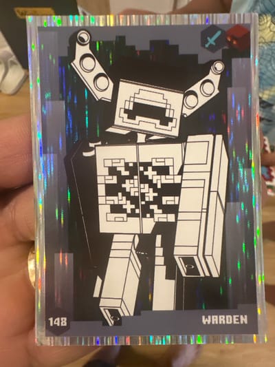 A person's hand is holding a holographic trading card featuring a stylized, pixelated robot character. The character, referred to as "Warden" by the text on the card, is predominantly black and white with a blocky design. The card itself has a holographic border with a rainbow effect. The background of the card depicts a dark, abstract city-like environment with vertical streaks of light. In the upper right corner, there is a small blue sword icon and a red block. The number "148" is visible in the lower left corner of the card. The hand holding the card has a gold ring on one finger. The environment outside the card is blurred, but appears to be an indoor setting with wooden surfaces. There is no information about the city of Mainburg, Germany in the image.