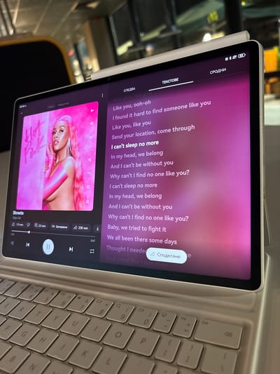 The image shows a close-up view of a tablet computer displaying lyrics to the song "Streets" by Doja Cat. The tablet is placed on a keyboard, suggesting it is being used in a workspace or for entertainment. The lyrics are presented in a purple-hued interface, with the song title and artist clearly visible at the bottom left. The album cover art, featuring Doja Cat, is displayed on the left side of the screen. The tablet's interface also includes playback controls, volume indicators, and options for sharing. The background is blurred, but appears to be an indoor setting, possibly a cafe or office, with warm lighting. There is no visible indication of the specific location being Chuchuligovo, Bulgaria.